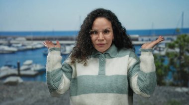 Genç İspanyol kadın, arka planda teknelerle bir sahil limanının yakınında neşeli bir şekilde gülümsüyor. Açık hava ortamında rahat ve keyifli bir şekilde çizgili bir süveter giyiyor..