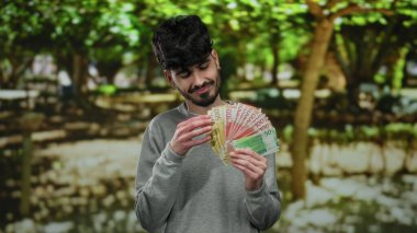 Elinde Norveç kronu banknotları tutan bir adam açık hava parkında gülümsüyor yemyeşil ve doğal aydınlatma ile huzurlu çevreyi vurguluyor..