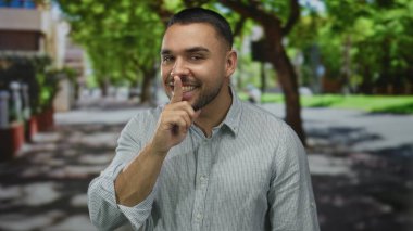 Şehir caddesinde, parlak güneş ışığında yapraklı ağaçların altında sessizlik için parmaklarını dudaklarına koyan adam. Gizlilik..