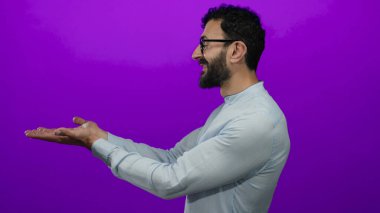 Gözlüklü İspanyol bir adam canlı mor bir arkaplan karşısında el kol hareketi yapıyor, bir sunum veya ürün sergiliyor davetkar bir gülümseme ve gündelik stil ile.