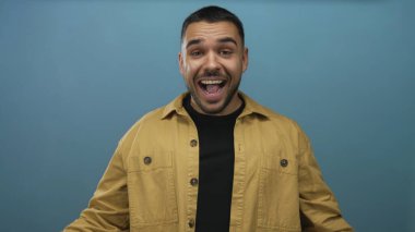 Hardal ceketli genç İspanyol adam mavi stüdyo duvarına karşı elleri ile gözlerini kapar hüzünlü poz; neşe dolu kaprisli kaprisli neşe.