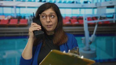 Orta yaşlı bir kadın yolcu gemisinde havuz kenarında siyah bir cep telefonu tutuyor..