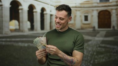 Açık üniversite ortamında Amerikan doları tutan genç bir adam, eski bir üniversite avlusunda renkli dövmeler sergilerken mutluluk ve zenginlik sergiliyor..