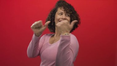 Stüdyoda pembe tişört giyen İspanyol kadın başparmağıyla göğsünü işaret ediyor; güven güçlendirme pozitifliği.