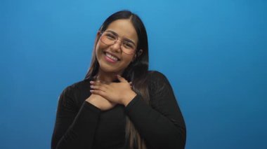 Mavi arka planda kollarını açmış gülümseyen genç bir kadın, gözlük takıyor ve siyah bir bluz giyiyor, izole edilmiş bir stüdyoda neşe ve açıklığın somutlaştırıcısı..