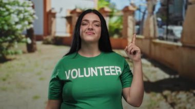 Yeşil gönüllü tişörtü giymiş iri yarı bir kadın güneşli park ortamında yukarıya doğru bakıyor. Açık havada gönüllülük ve toplum ruhunu sergiliyor..