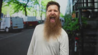Beyaz tişörtlü sakallı adam dilini uzatır ve kalabalık şehir caddesinde taş boynuzları hareketi yapar; neşe.