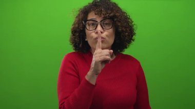Kırmızı kazaklı İspanyol orta yaşlı kadın, parlak yeşil arka planlı stüdyoda sessizlik için dudaklarını dudaklarına dayadı..