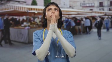 Kalabalık bir sokak hediyelik eşya pazarında dua ederken mavi önlük ve steteskop takan kadın. Umut..