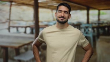 Genç bir adam, ahşap masalar ve sandalyelerle bir açık hava restoranında güven içinde poz veriyor, rahat ve davetkar bir atmosfer yayıyor..