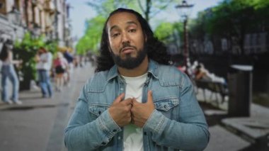 Elleri göğsünde ve açık avuç içleri kalabalık bir caddede omuz silken, kot ceket ve beyaz tişört giyen, sakalı görünen ve yayalar arkasında belirsizlikler olan bir adam..