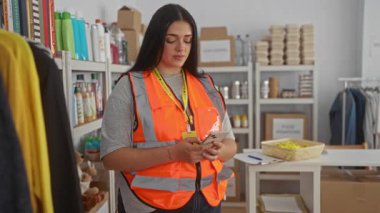 Bağış merkezinde gönüllü olarak çalışan bir kadın. Etrafı ürün ve kıyafetlerle çevriliyken telefonu kontrol ediyor. Kapalı bir hayır kurumunda parlak bir yelek giyiyor..