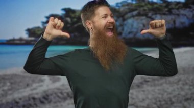 Deniz kenarındaki sahilde uzun sakallı, yeşil tişörtlü, gülen ve baş parmaklarını göğsüne gösteren bir adam..