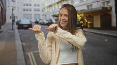 Kadın her iki başparmağını da geriye doğru uzattı, kesikli üst ve parmağında görünen yüzükle gülümsedi; oyuncu özgüven.