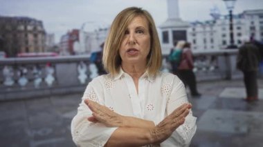 Kadın, şehir caddesinde x jestinde, bulanık yayalarla tırabzan yaparak önkollarını çaprazlıyor..