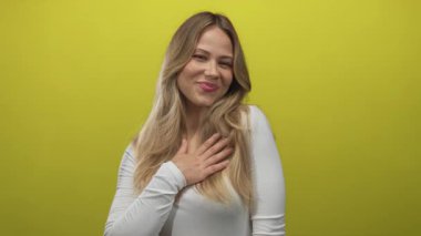 Sarışın beyaz kadın, fotoğraf stüdyosunda yumuşak bir gülümsemeyle elini göğsüne koyar; minnettarlık samimiyeti takdir.