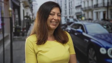 Kalabalık bir caddede, park etmiş arabaları ve binaları olan sarı bluzlu, görünür yüzüyle gülümseyen İspanyol kadın. Mutluluk..