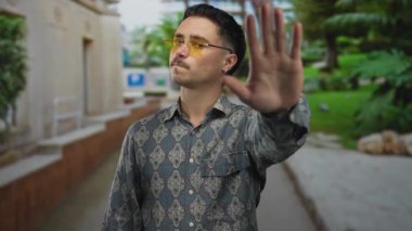 Young hispanic man wearing patterned shirt and yellow glasses gesturing stop in a lush park setting with greenery and pathways visible behind him outdoors