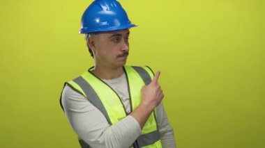 Yansıtıcı yelek giyen ve kasketi parmağıyla izole edilmiş sarı arka plan duvarını işaret eden İspanyol genç adam, güvenlik odaklı bir inşaat konseptini gösteriyor..