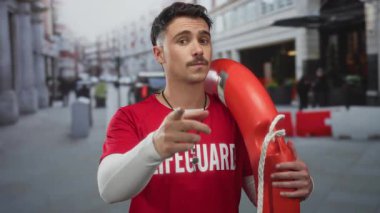 Kırmızı tişört giyen genç İspanyol cankurtaran kalabalık şehir caddesinde dükkan ve arka planda insanlar varken şamandıra tutuyor.