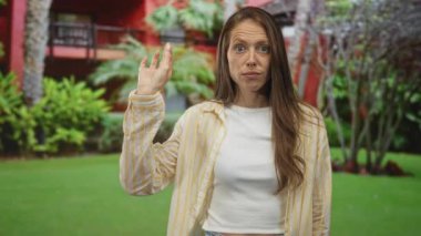 Sağ elini kaldıran genç bir kadın kırmızı bir bina ve dışarıda palmiye ağaçları ile peyzaj bir çimenlik yanında sorgulama jesti; belirsizlik.