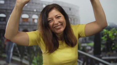 Orta yaşlı İspanyol kadın sarı tişörtlü gülümseyen kadim kolezyum binasından önce el çırpıyor; seyahat heyecanı sevinci.