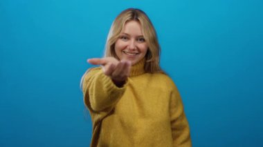 Sarı süveter giymiş, mavi arka planda gülümseyen bir kadın, sarı saçlı, beyaz tenli, davetkar bir şekilde el ele tutuşan, neşeli ve sakin bir ortam..