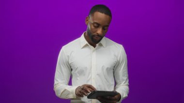 Genç Afro-Amerikalı bir adam tableti parmaklarıyla tıklatıyor, beyaz gömlek giyiyor ve mor arka planda stüdyoya bakıyor..