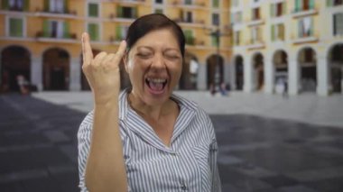 Kaya boynuzları yapan kadın, sarı bina cephesinde tarihi kemerleri olan güneşli bir şehir caddesinde el hareketi yapıyor..