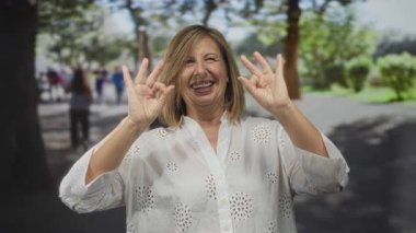 Orta yaşlı bir kadın geniş bir gülümseme takınıyor ve yeşil yapraklı orman parkı ağaçlarının arasına işaret koyuyor; onay sükuneti.