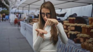 Kadın, boş el ve işaret hareketi sunarken, sokak pazarındaki tezgahlarda el kaldırmayı işaret ediyor; şüphecilik.