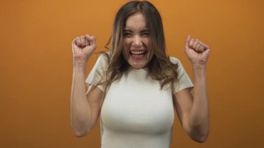 Beyaz gömlekli bir kadın kendine güvenerek gülümsüyor ve canlı turuncu stüdyoda yumruklarını sallıyor..
