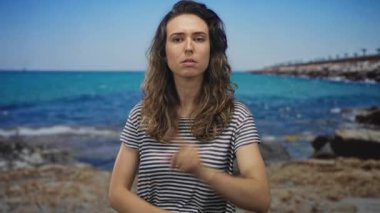 Güneşli çizgili gömlekli kadın deniz kenarındaki kayalık rıhtımın yakınındaki sokakta kameraya işaret ediyor..