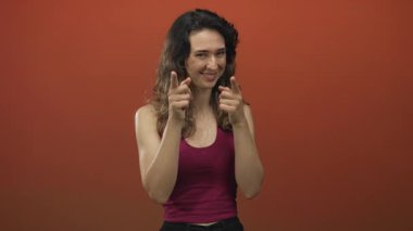 Genç İspanyol kadın parmaklarını turuncu stüdyoda şen şakrak sırıtarak ve parmak tabancası hareketiyle öne doğru gösteriyor..