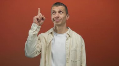 Kırmızı duvara yaslanmış bej gömlekli bir adam, düşünceli bir ifadeyle yukarıya doğru bakıyor. İzole edilmiş bir alanda fikri ya da ilhamı simgeliyor..