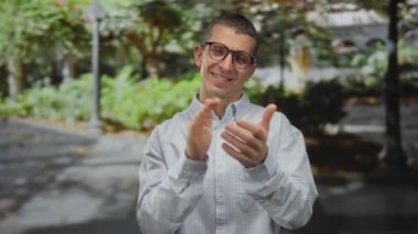 Beyaz bir adam, yeşil ve benekli güneş ışığıyla dolu bir parkta el çırpıyor, gözlük takıyor ve çizgili bir gömlek giyiyor, bir başarı duygusu ve pozitif enerjiyi simgeliyor..