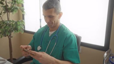 Steteskoplu yeşil üniformalı beyaz erkek doktor hastane odasında akıllı telefon kullanıyor. İçi bitki ve telefon dolu..