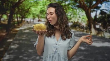 Yeşil ağaçlarla kaplı bir sokakta cam kaseden çıtır patates cipsi yiyen bir kadın, zevkten zevk alır..
