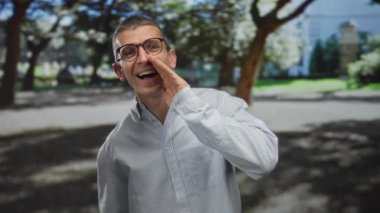 Güneşli bir parkta yüksek sesle bağıran, gözlüklü ve çizgili tişörtlü bir adam. Ağaçlar etrafını gölgeleyecek, açık havada enerji taşıyacak..