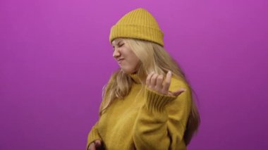 Genç sarışın kadın hardal süveter giyip pembe arka planda gitar taklidi yapıyor..