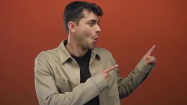 Adam arka planda kameraya işaret ederken parmağını kaldırıyor ve turuncu giyen genç Latin erkek şaşırma hareketi gösteriyor..