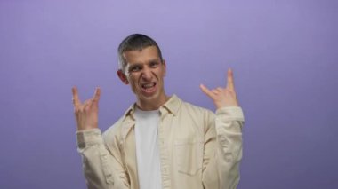 İki eliyle kaya jesti yapan, canlı mor bir arka plana karşı heyecanını ifade eden, bej gömlek ve beyaz tişört giyen, neşeli ve anlamlı duyguları vurgulayan bir adam..