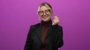 Pembe arka planda kalp işareti yapan bir kadın stüdyoda kendine güvenen bir şekilde gülümsüyor ve siyah ceket giyiyor.