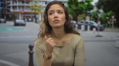 Saati olan kadın şehir caddesinde binalar ve arka planda yeşillik ile düşünceli görünüyor. Duygularını ve düşüncelerini aktarıyor..