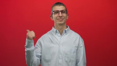 Beyaz bir adam gülümsüyor ve başparmağıyla işaret ediyor kırmızı arka planda, izole edilmiş, günlük gömlek giyiyor, stüdyo ortamında kendine güvenini ve pozitifliğini ifade ediyor..