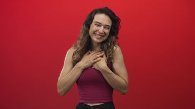 Kırmızı stüdyo ortamında elleri göğsünde ve kolları açık gülümseyen bir kadın..