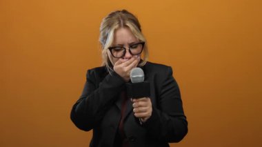 İzole edilmiş turuncu arka planda mikrofon tutan kadın muhabir siyah gözlük ve ceketle duygularını ifade ediyor.