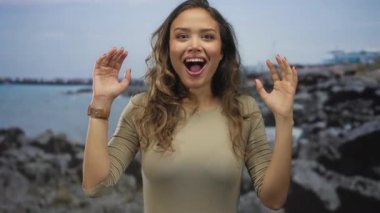 Heyecanlı yüzlü bir kadın bulutlu bir gökyüzünün altında kayalık bir sahil kenarında duruyor. Elleriyle doğanın güzelliğini ve duygularını gösteriyor..