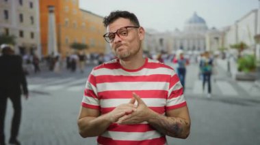 Vatikan şehir meydanında gözlüklü genç adam kırmızı çizgili tişört giyiyor. İnsanlar ve şehir geçmişi güneşli bir günde sokak modası ve modern dövme sergiliyor..