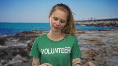 Kumsaldaki sarışın gönüllüye terini silen, güneşli okyanus göğünün altında yeşil tişört giyen kayalık önplanlı ve yorgun görünen bir kadının portresi..